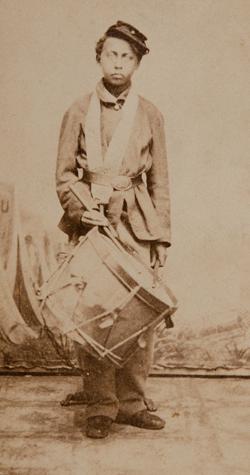 <p>A black and white photograph of a young, 一个黑人男孩，头戴一顶帽子，身穿一件大军大衣，腰间系着一条腰带，衬着白色的背景. 他一手拿着一只鸡腿, 其中一个靠在一个挂在脖子上的大军鼓上.</p>