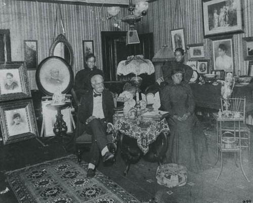 <p>Black and white photograph with a white border depicting a room with striped wallpaper and decorated with several portraits with four people in the center, 围绕着桌子和台灯. 一个穿着西装、跷着二郎腿的黑人老人坐在桌子对面，对面是一个穿黑裙子的黑人妇女. 另外两个穿着黑色连衣裙的黑人妇女站在桌子两边的两个人后面.</p>